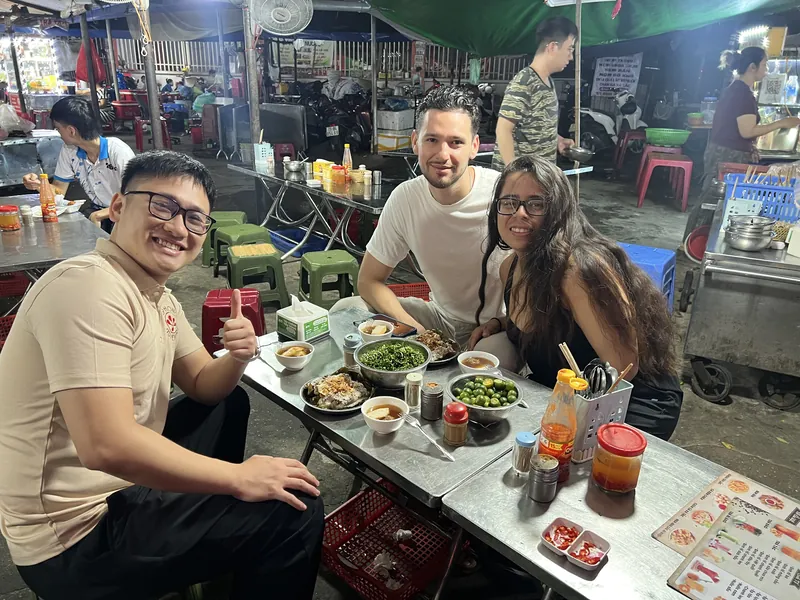 Guide and foreign couple eating banh beo at a busy Haiphong sidewalk stall at night